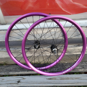 A pair of 24-inch TNT M80X purple anodized BMX wheels with black spokes and brass nipples, displayed on a wooden surface.