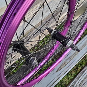 A pair of 24-inch TNT M80X purple anodized BMX wheels with black spokes and brass nipples, displayed on a wooden surface.