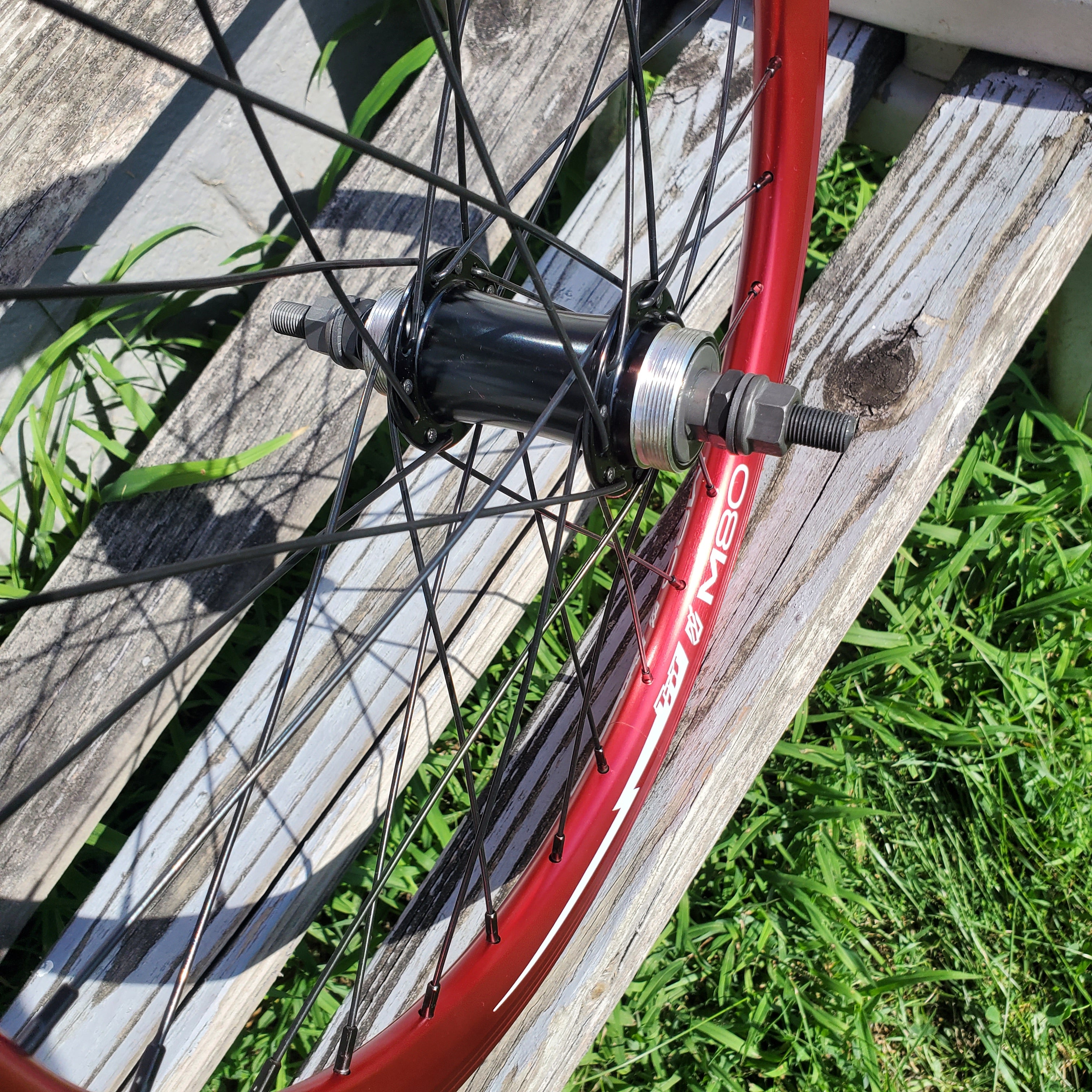 Close-up of a bicycle wheel with red rim and black spokes on a wooden surface with grass in the background.