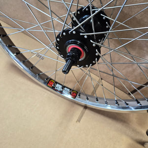 Close-up of a bicycle wheel hub and rim with silver spokes, a black hub, and a small red ring detail, photographed against a tan cardboard background.