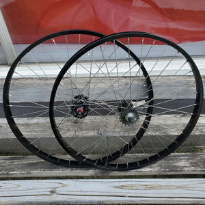A pair of 26-inch black BMX wheels with coaster brake bands, hubs, and spoke nipples visible.