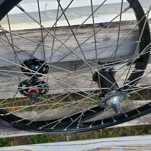 A pair of 26-inch black BMX wheels with coaster brake bands, hubs, and spoke nipples visible.